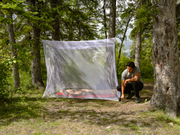 Young man hammering stakes into the ground to set up a white mesh tent surrounded by a forest.