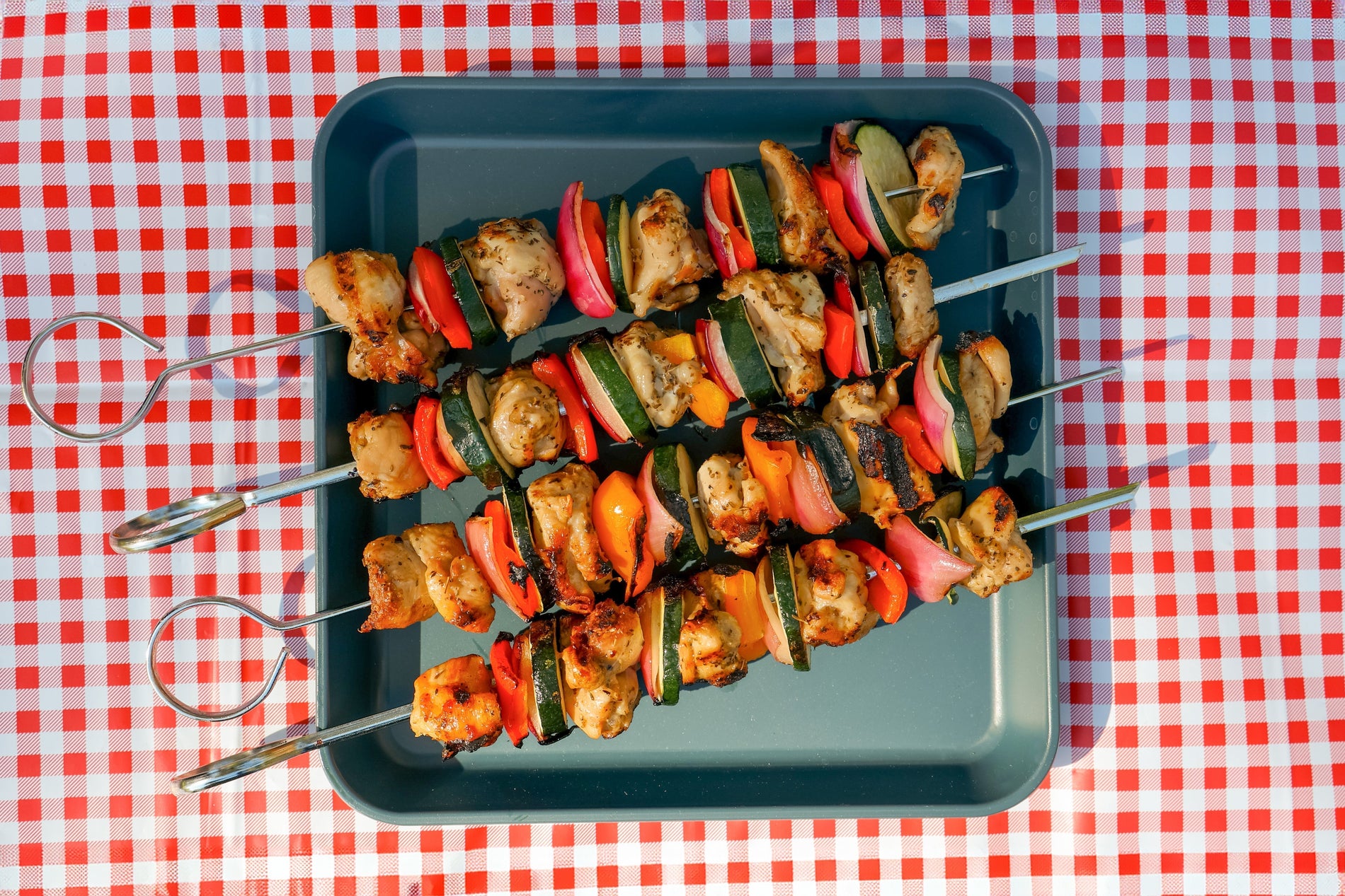 Four grilled chicken and vegetable kebabs on a blue plate placed on a red and white checkered tablecloth