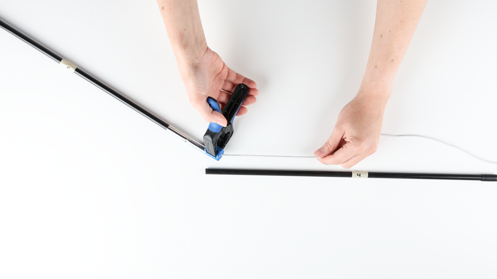 On a white table, a person's hands using a clamp to pinch a shock cord on the tip of the second to last section of a tent pole. 