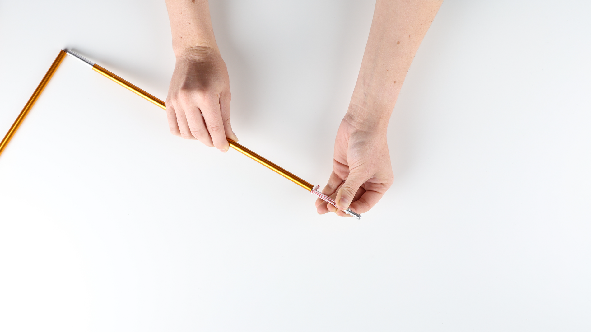 On a white table, a person's hands holding an Orange aluminum tent pole. One hand is pulling out the metal tabs and exposing the tip of the shock cord on one end of the pole.