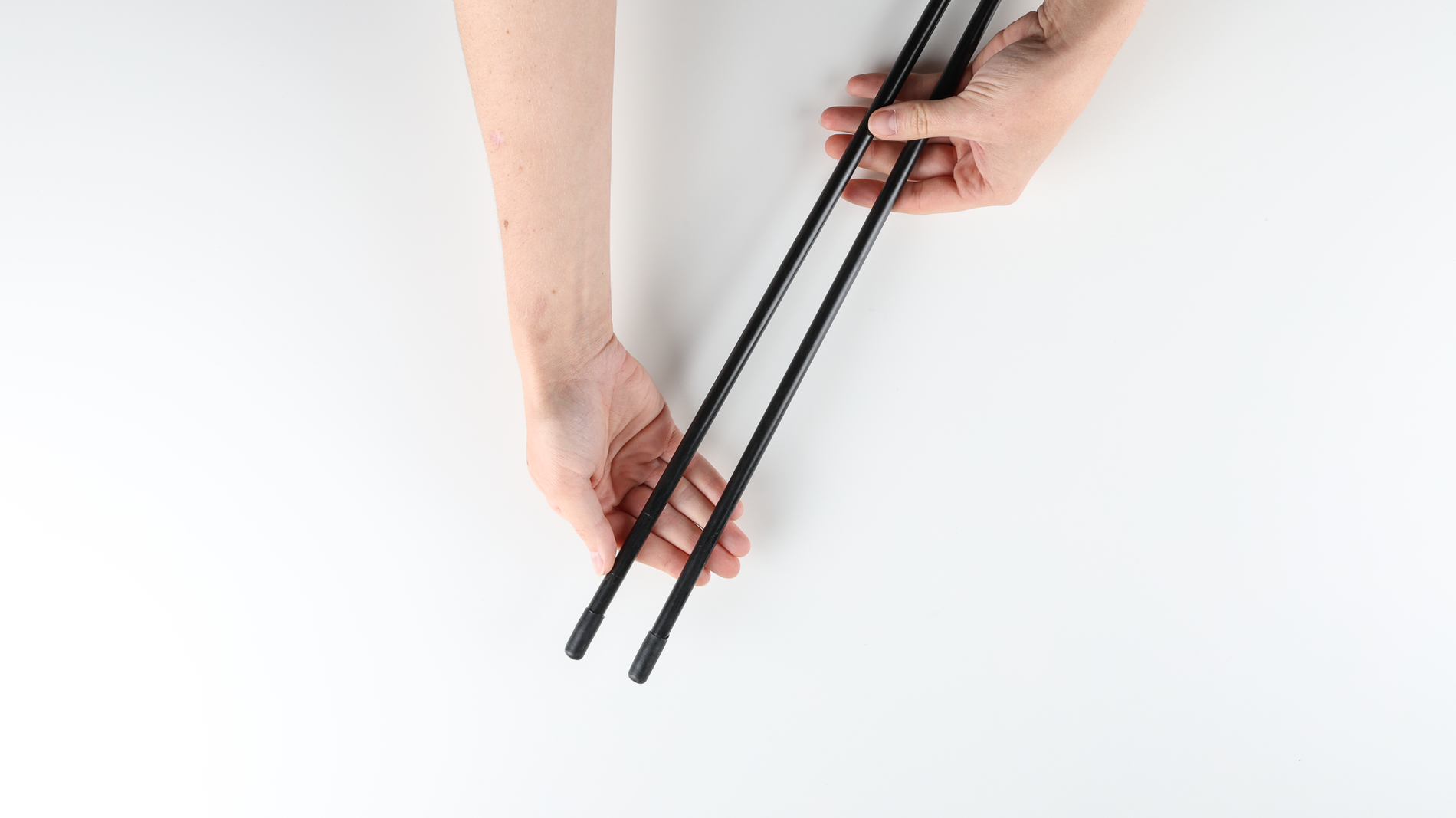 On a white table, a person's hands holding the two ends of a collapsed black fiberglass tent pole. The end tabs are made of plastic.