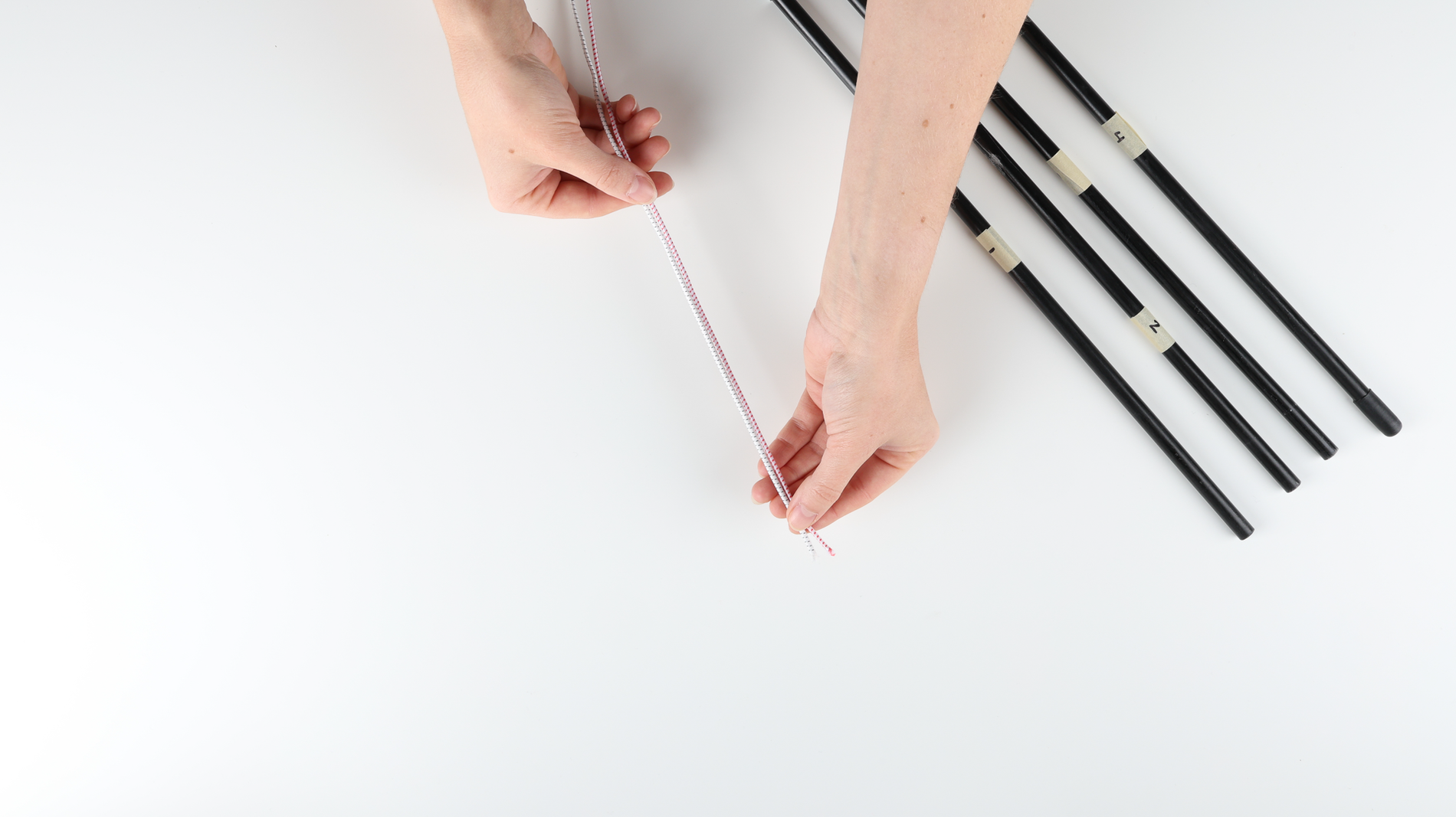 On a white table, a person's hands measuring an old shock cord (red and white color) to cut and match the length with a new shock cord (white color). To the right side are 4 black tent poles numbered with tape from no. 1 to 4