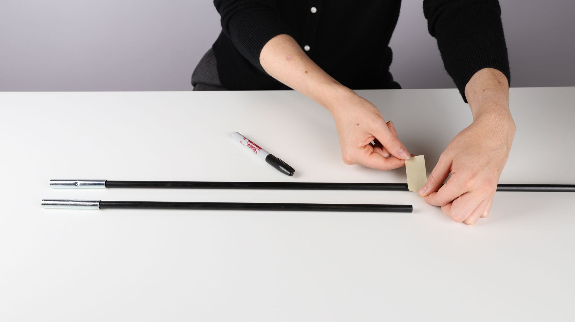 At a table, a person's hands are seen measuring the length of a shorter, broken tent pole. They are also marking with tape the longer Coghlan's replacement tent pole in preparation for cutting.