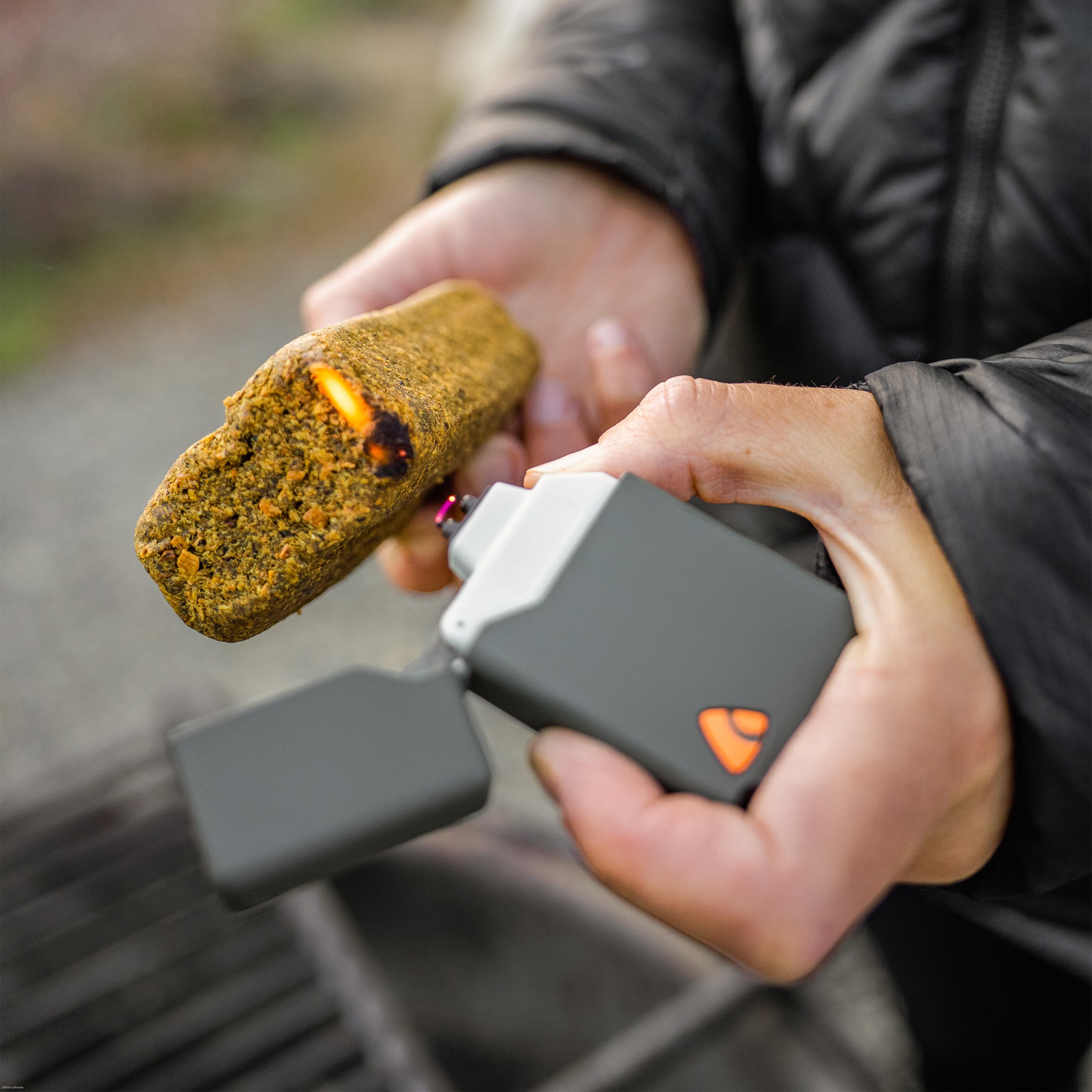 Close-up of hands using a grey plasma lighter to light the edge of a fire-starter over a firepit