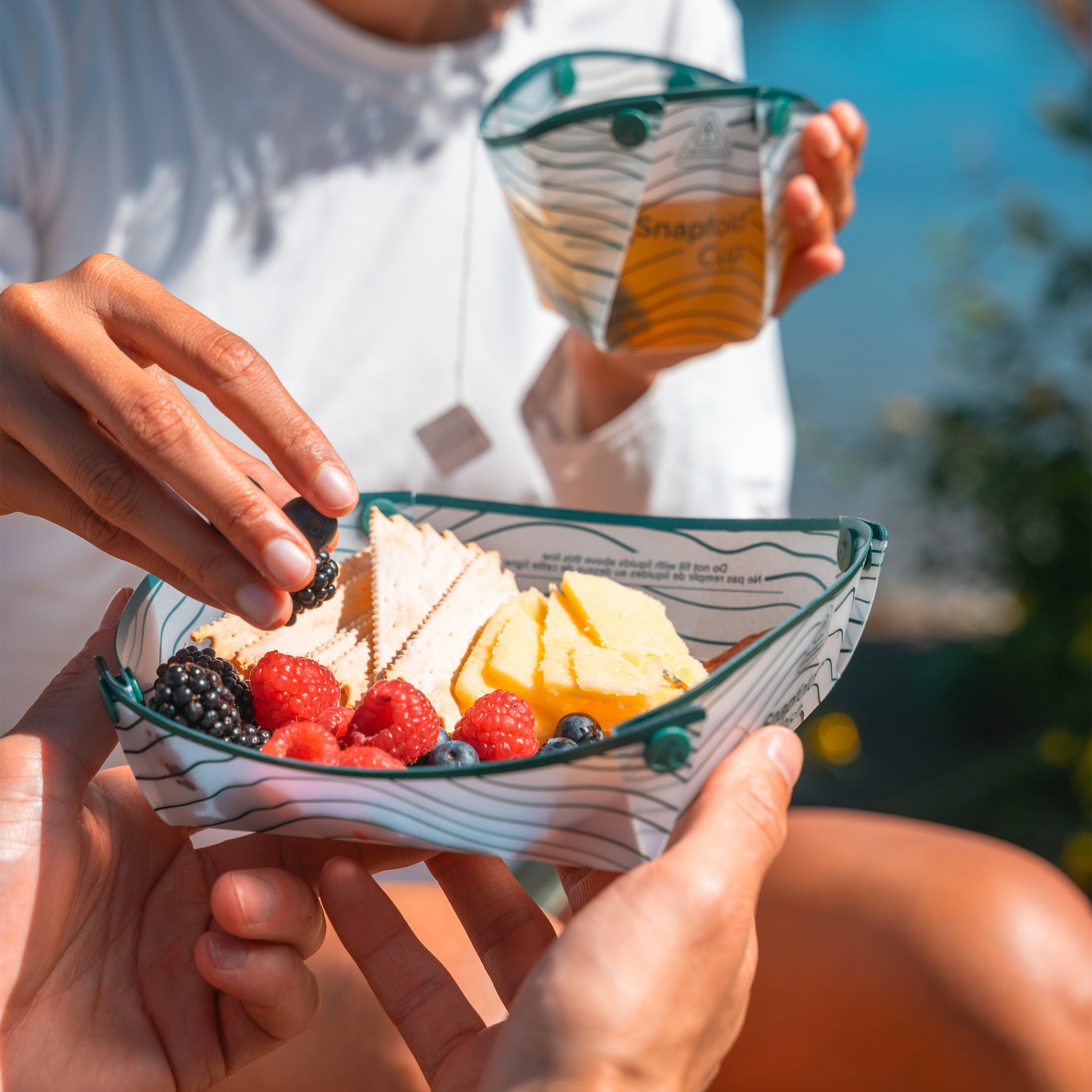 Close-up of hands taking berries and crackers out of a collapsible plastic dish while seated outside