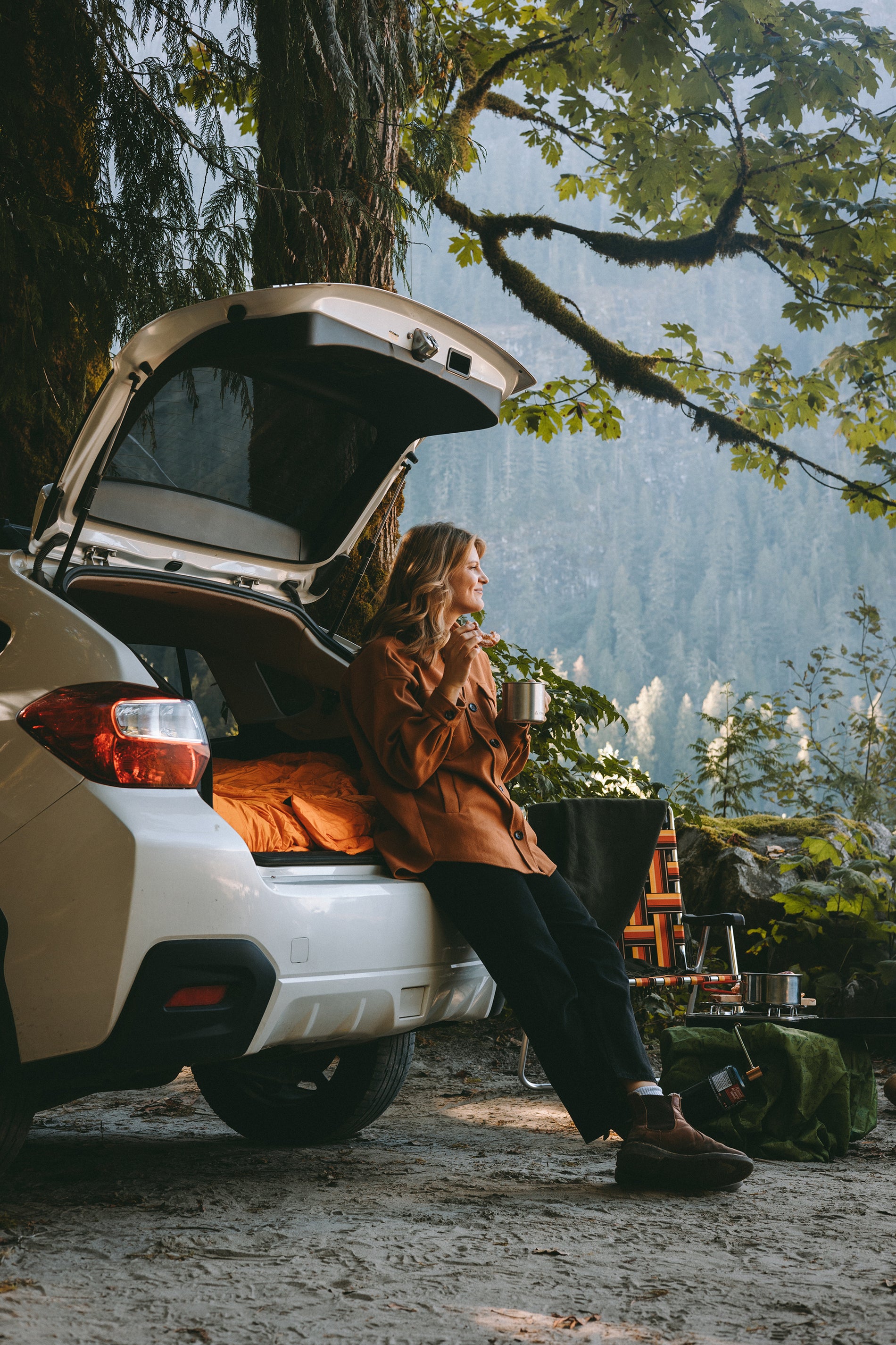 A woman leaning on the hitch of her car enjoying a cup of coffee at a campsite in the early morning