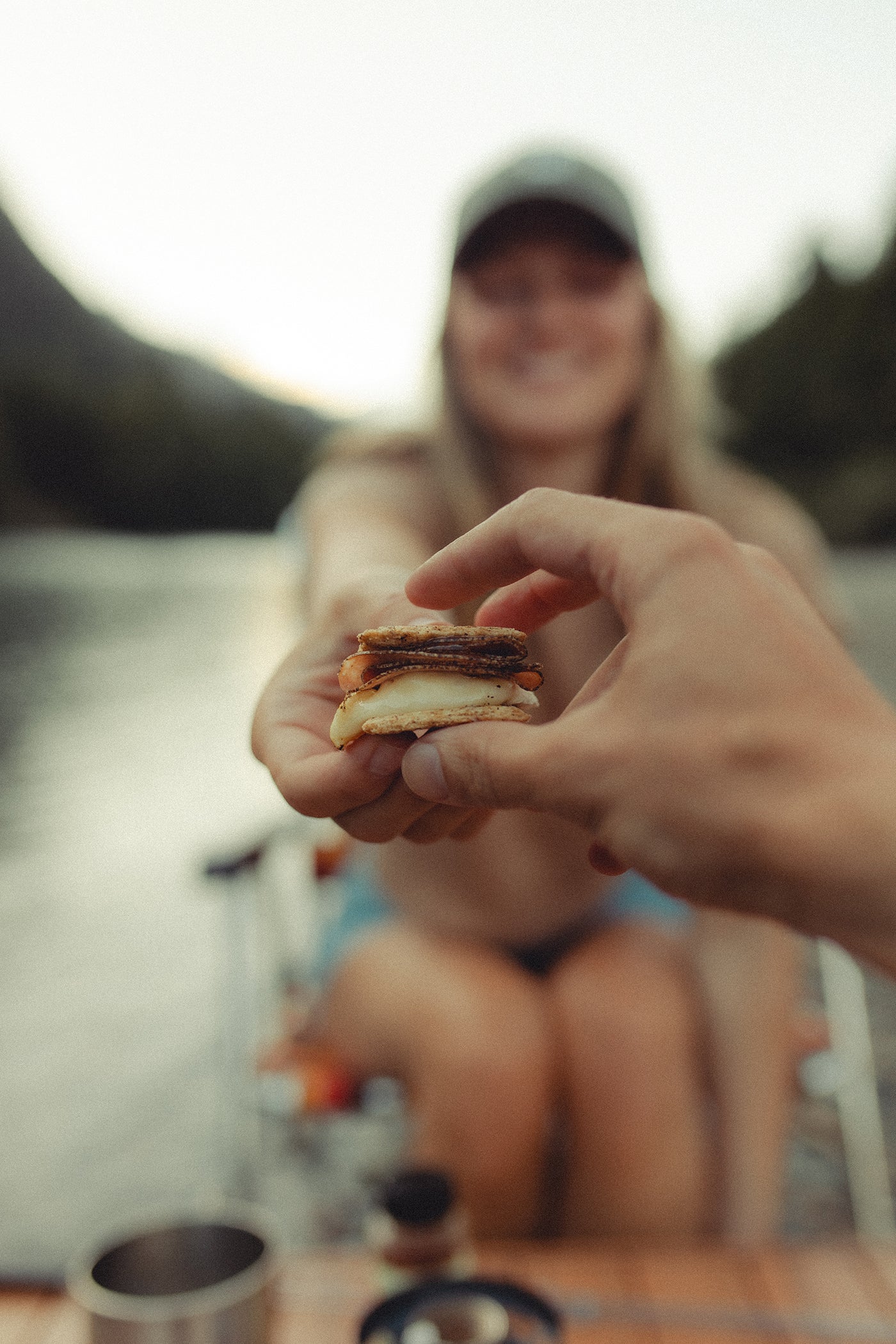 Person holding a piece of cheese and salami between crackers in front of a blurred outline of a woman sitting at a campsite