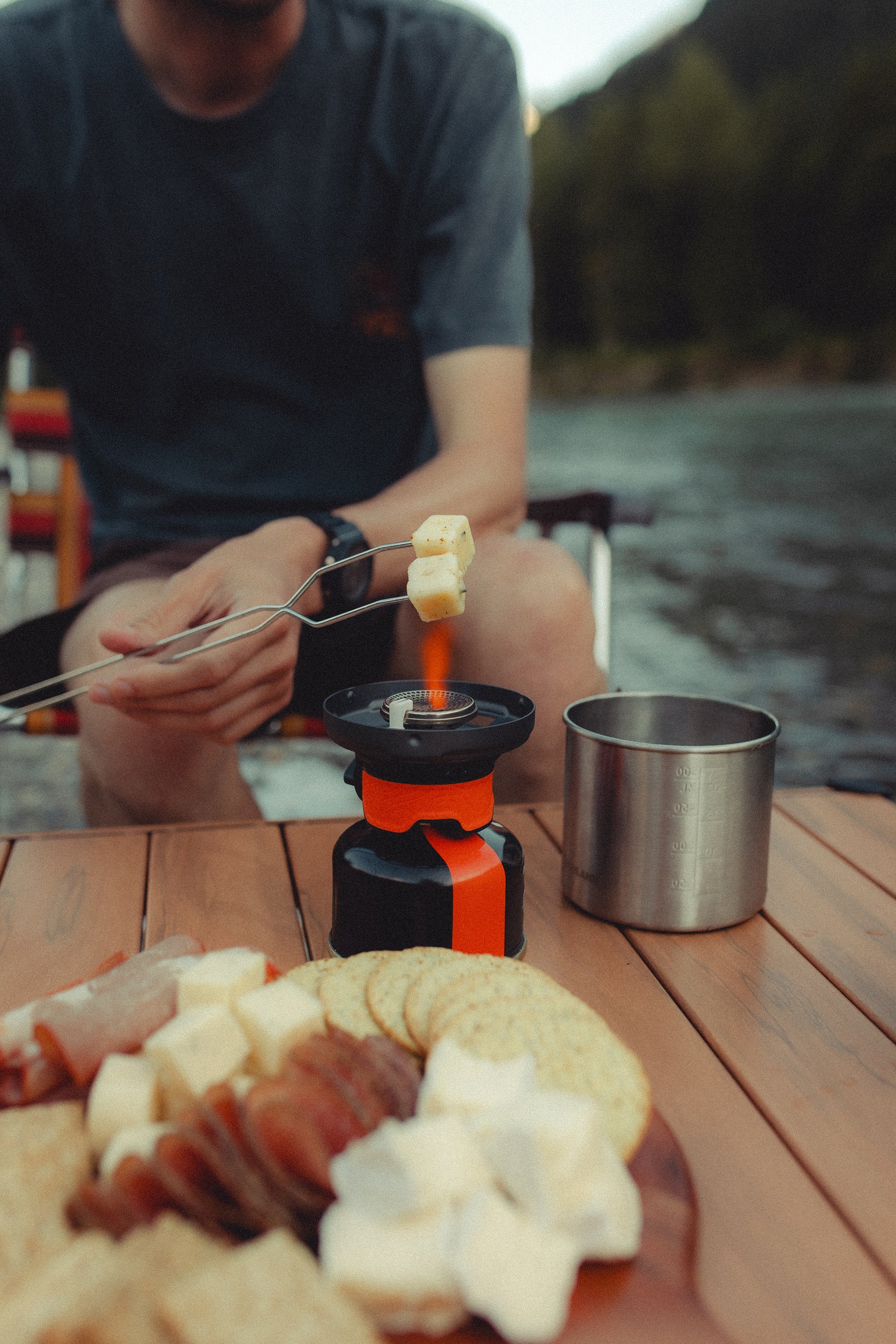 Close-up of a man's torso sitting at a portable table enjoying assorted meat, cheese and crackers while melting cheese over a portable stove