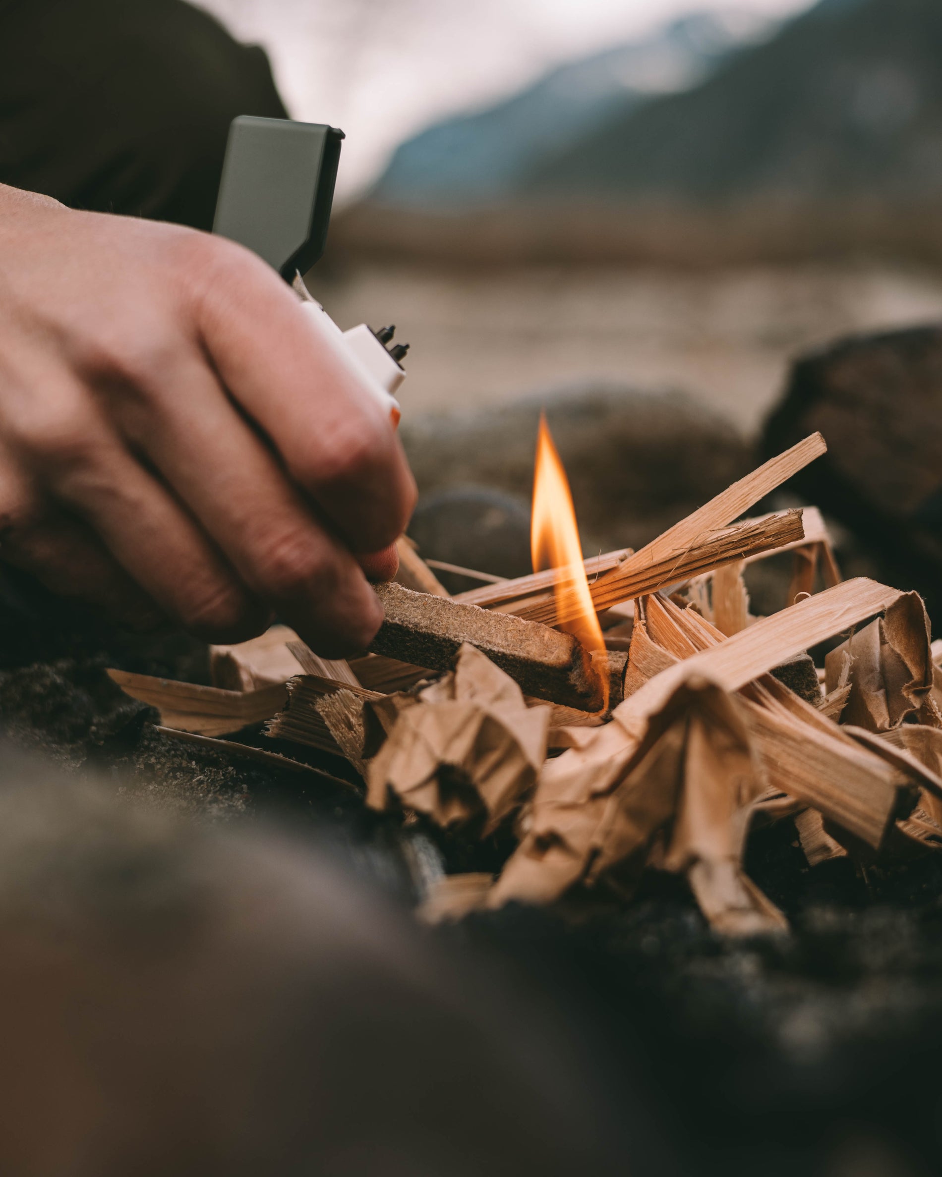 Close-up of a hand holding a plasma lighter to light a pile of tinder and kindling in a fire pit along a river shore