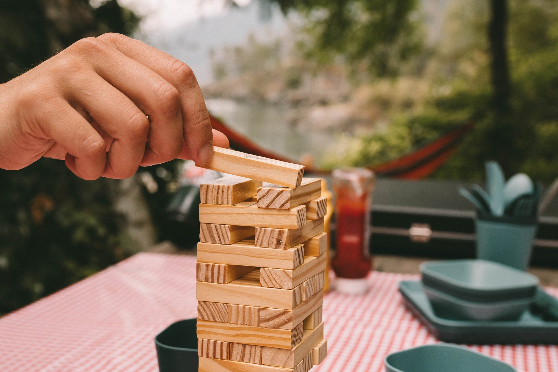 Hand carefully placing a block on top of tower of Jenga blocks with a blurred camping set-up in the background