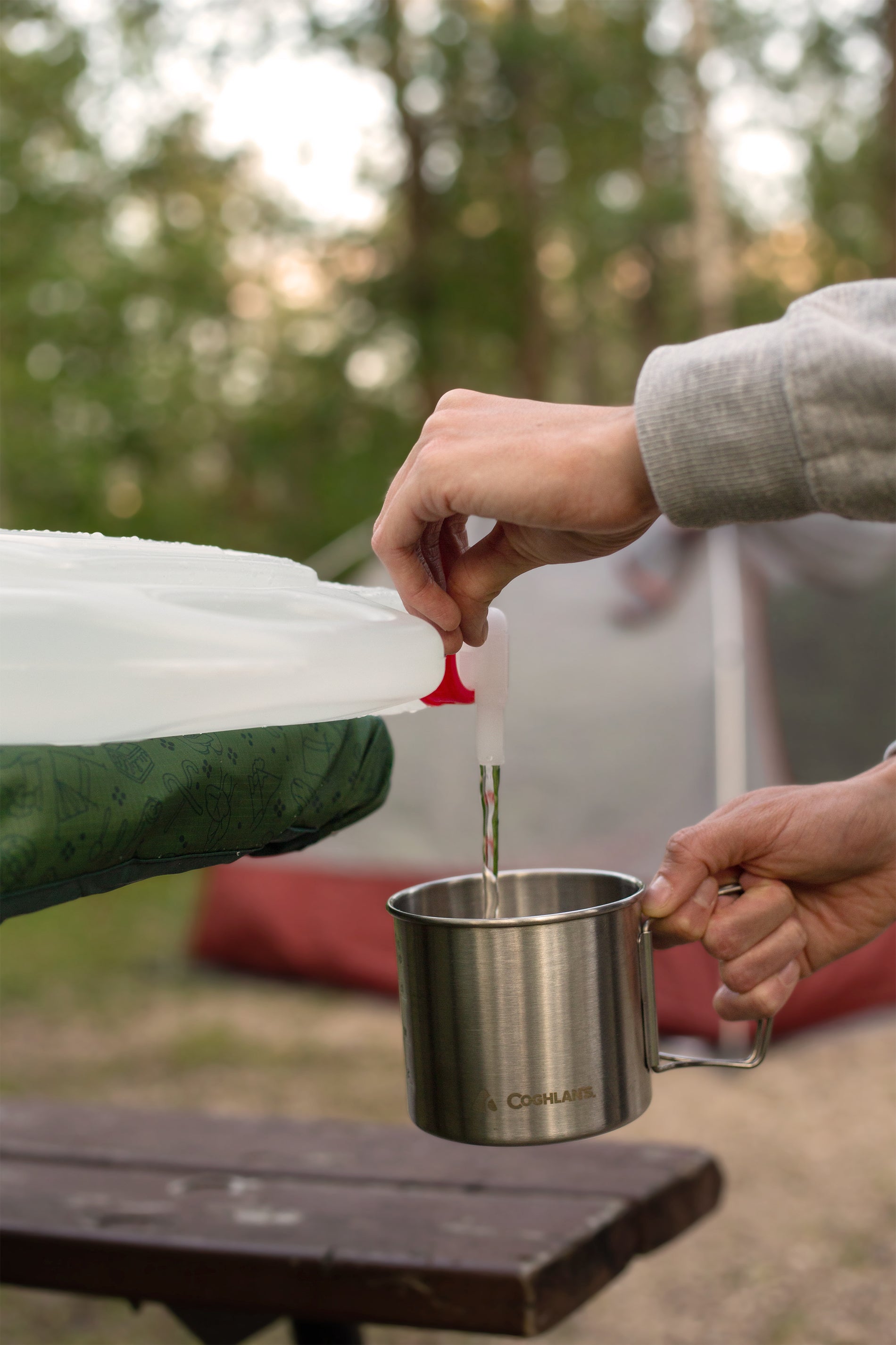 Close up of a person's hands filling a metal mug from a water container on a picnic bench