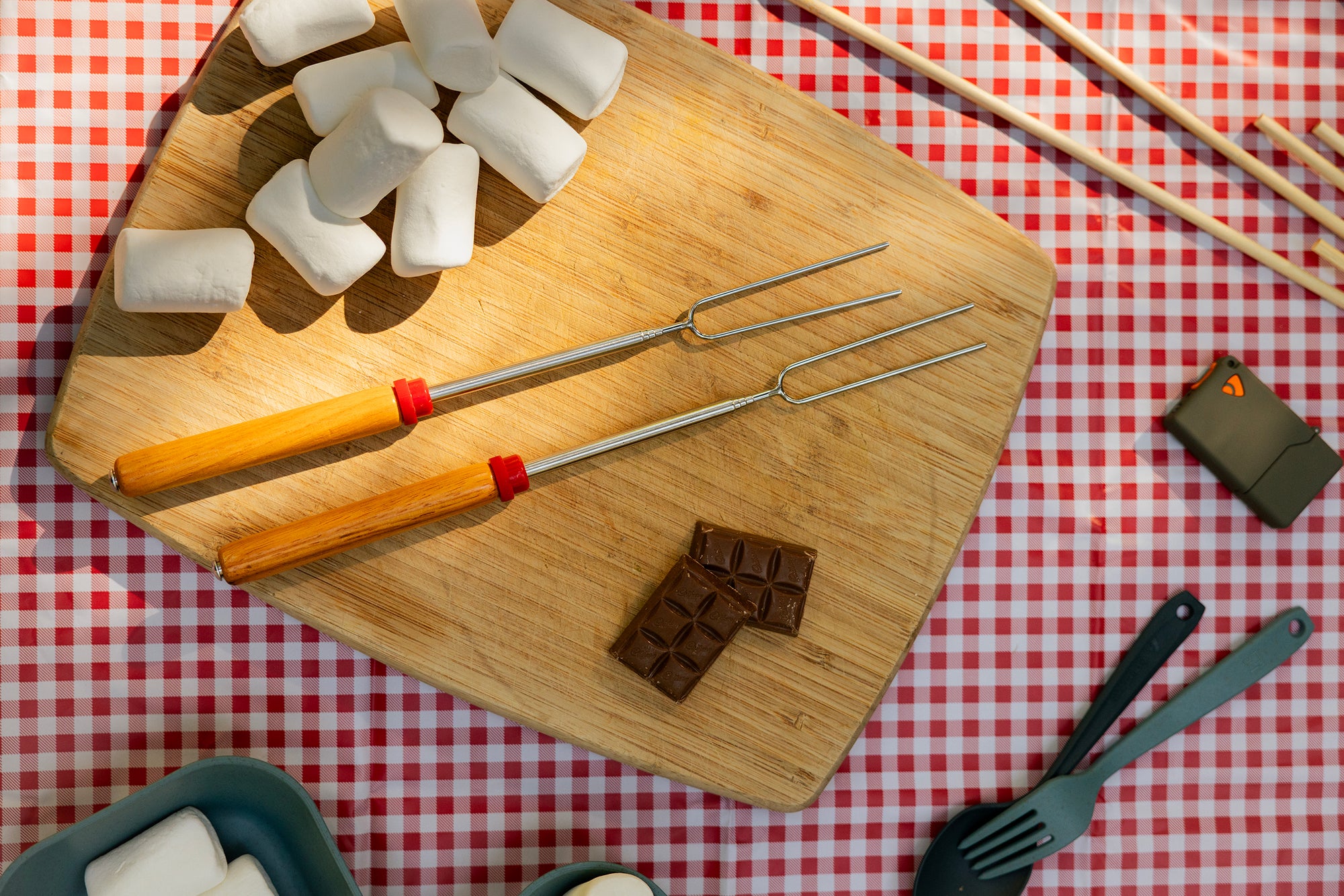 Wooden cutting board with marshmallows, chocolate, and skewers on a red and white checkered tablecloth.