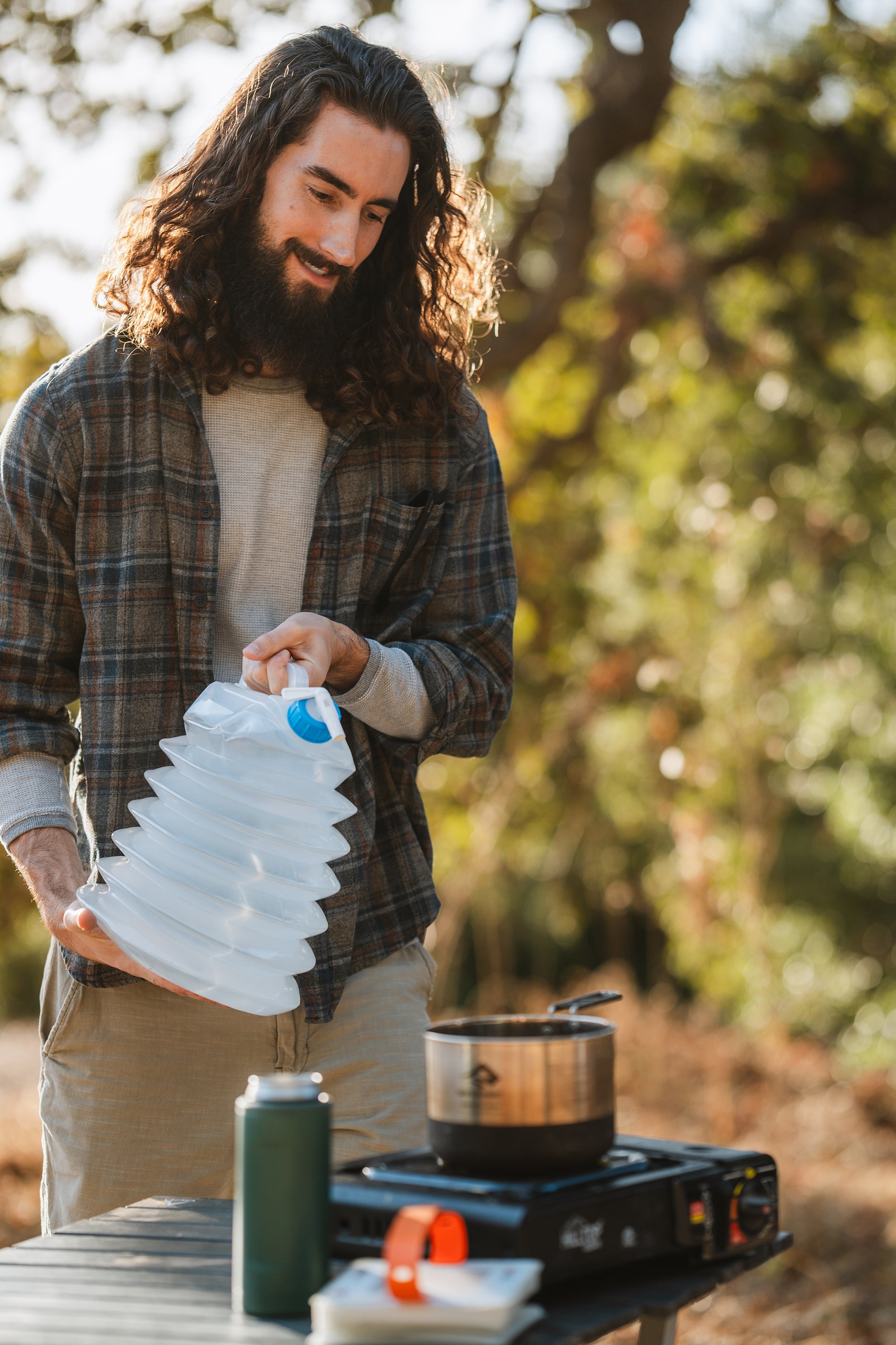 Young man with long hair and beard pouring water from a water container into a pot on a portable camp stove