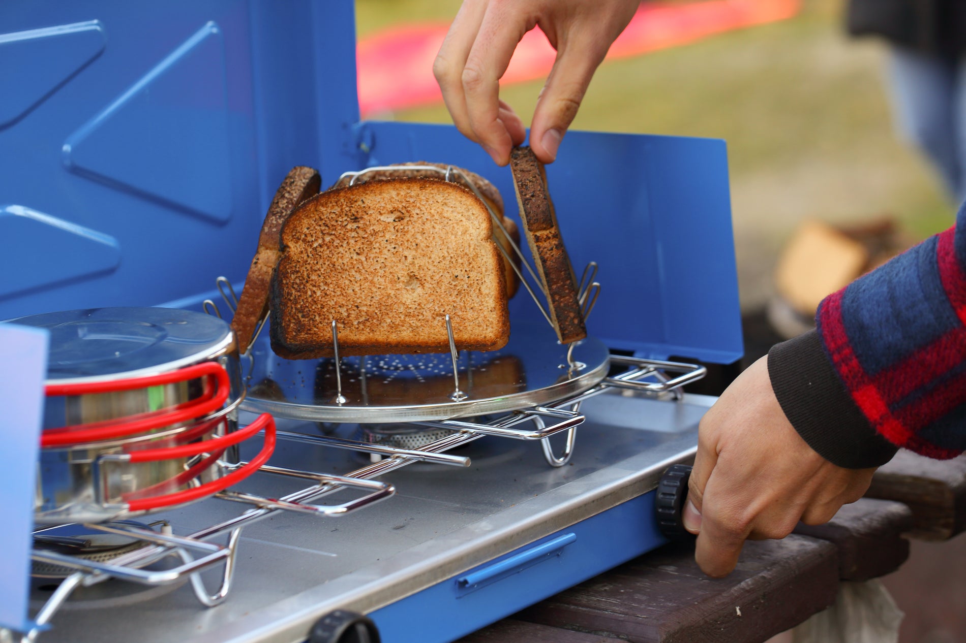 A person cooking toast on a picnic table using Coghlan's Camp Stove Toaster.