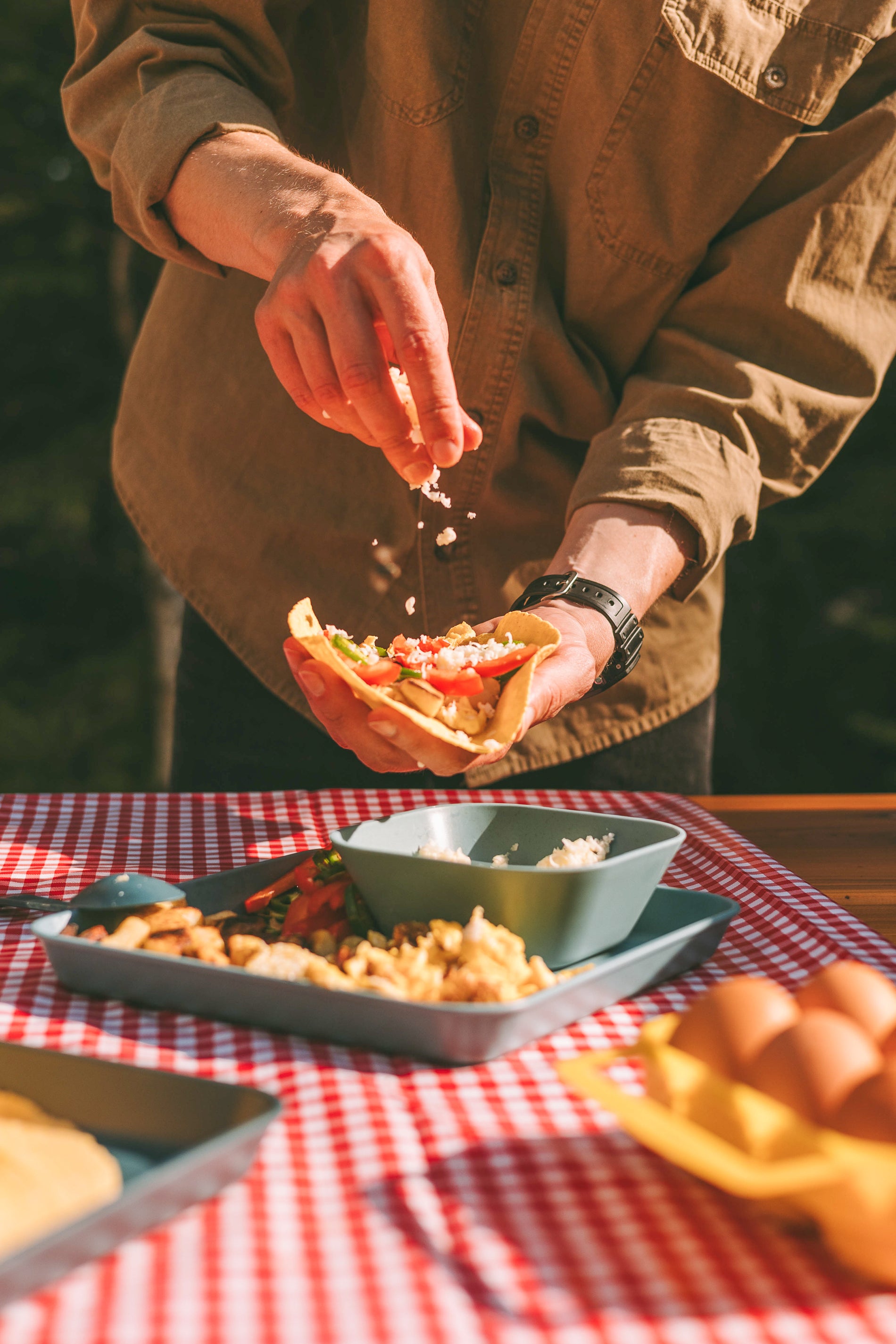 A person's hands holding a taco and garnishing it with cheese on a picnic table