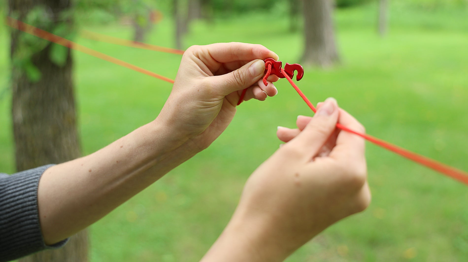 A person's hands looping a cord around a tree and using the anchor clip set's tensioner to tighten and secure the one end of the cord being hanged between two trees. Continuation of the previous step