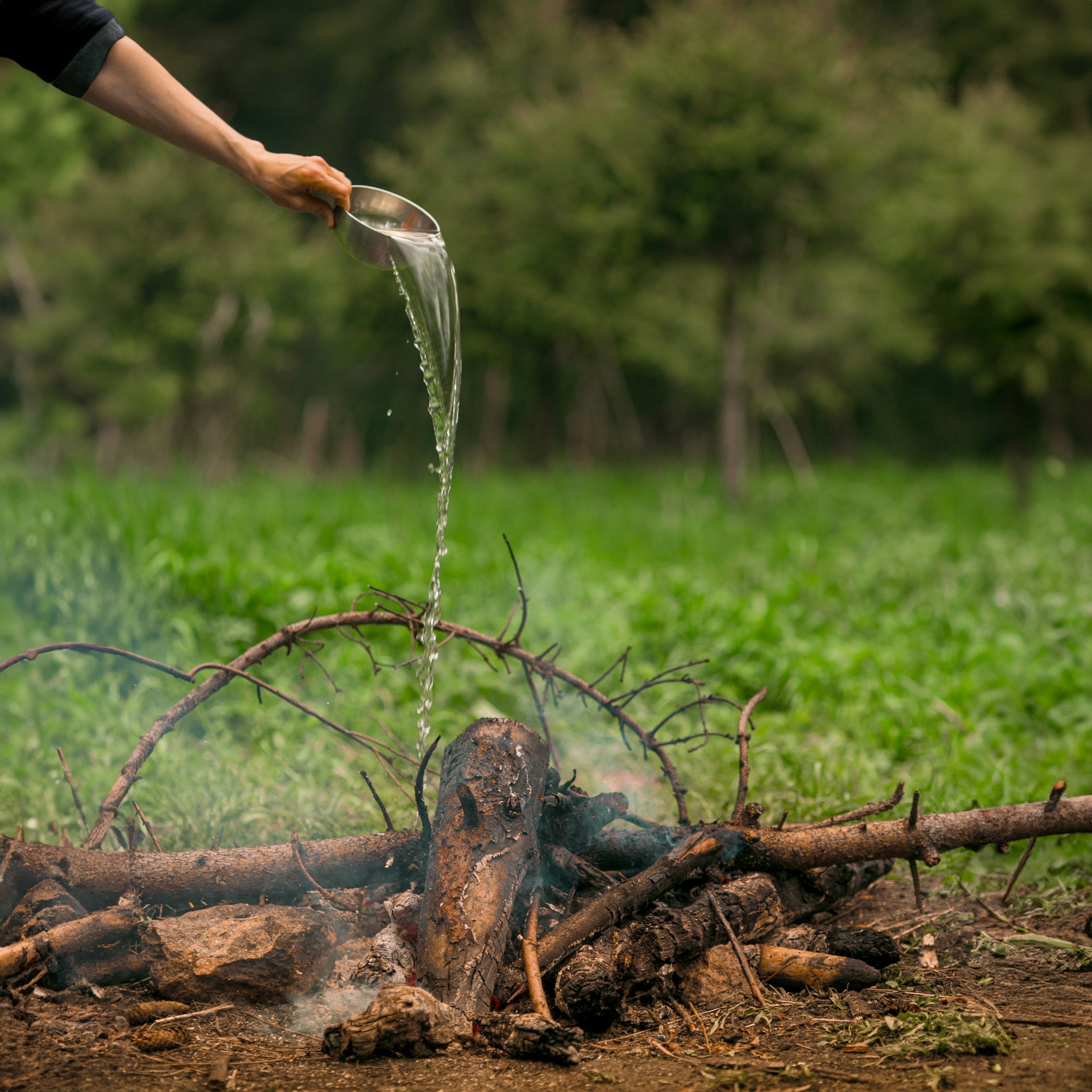 An outreached hand pouring a bowl of water over a fire at a campsite with smoke rising upwards