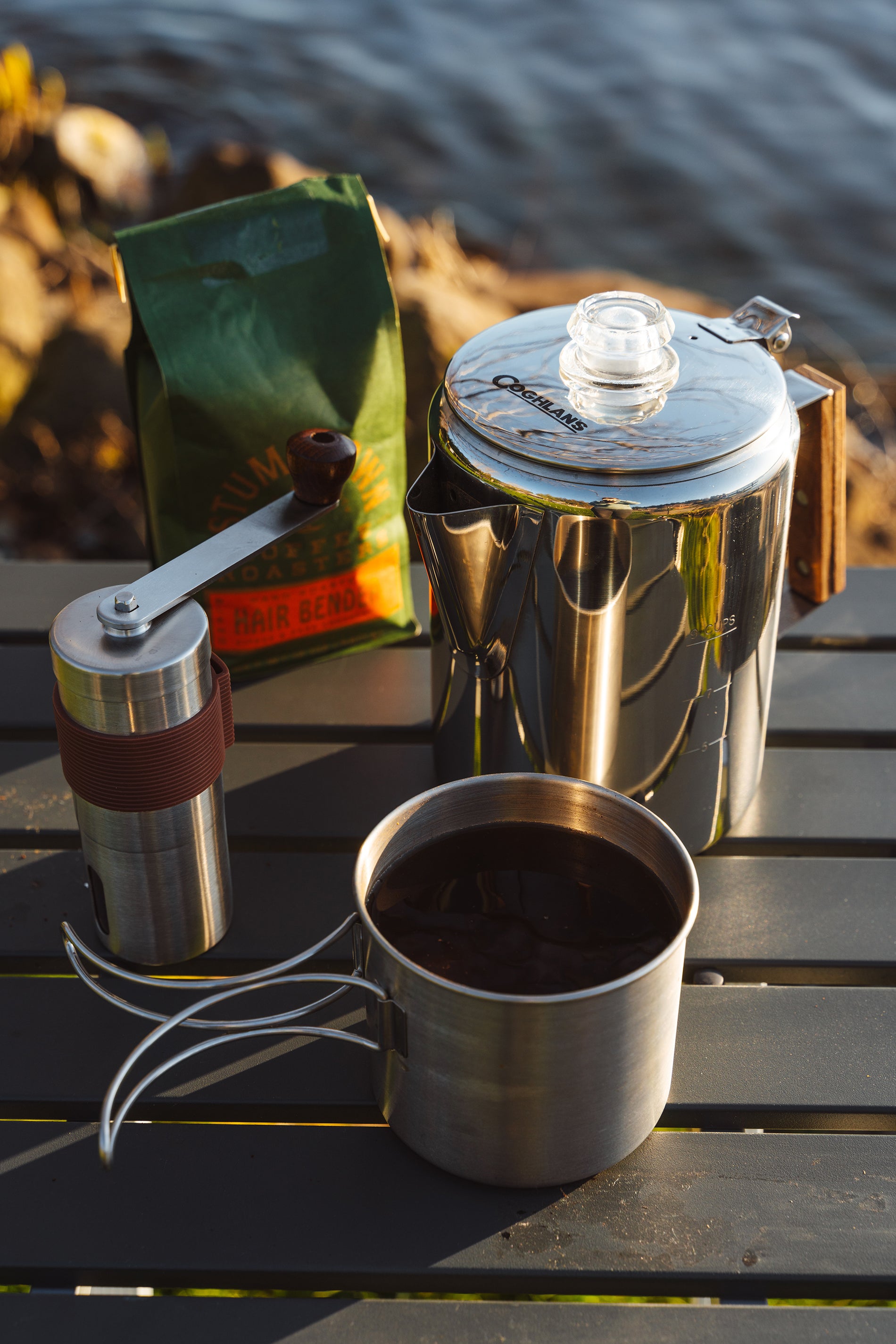 A camp mug, percolator coffee pot, coffee grounds and a coffee grinder on top of a picnic bench at a campsite