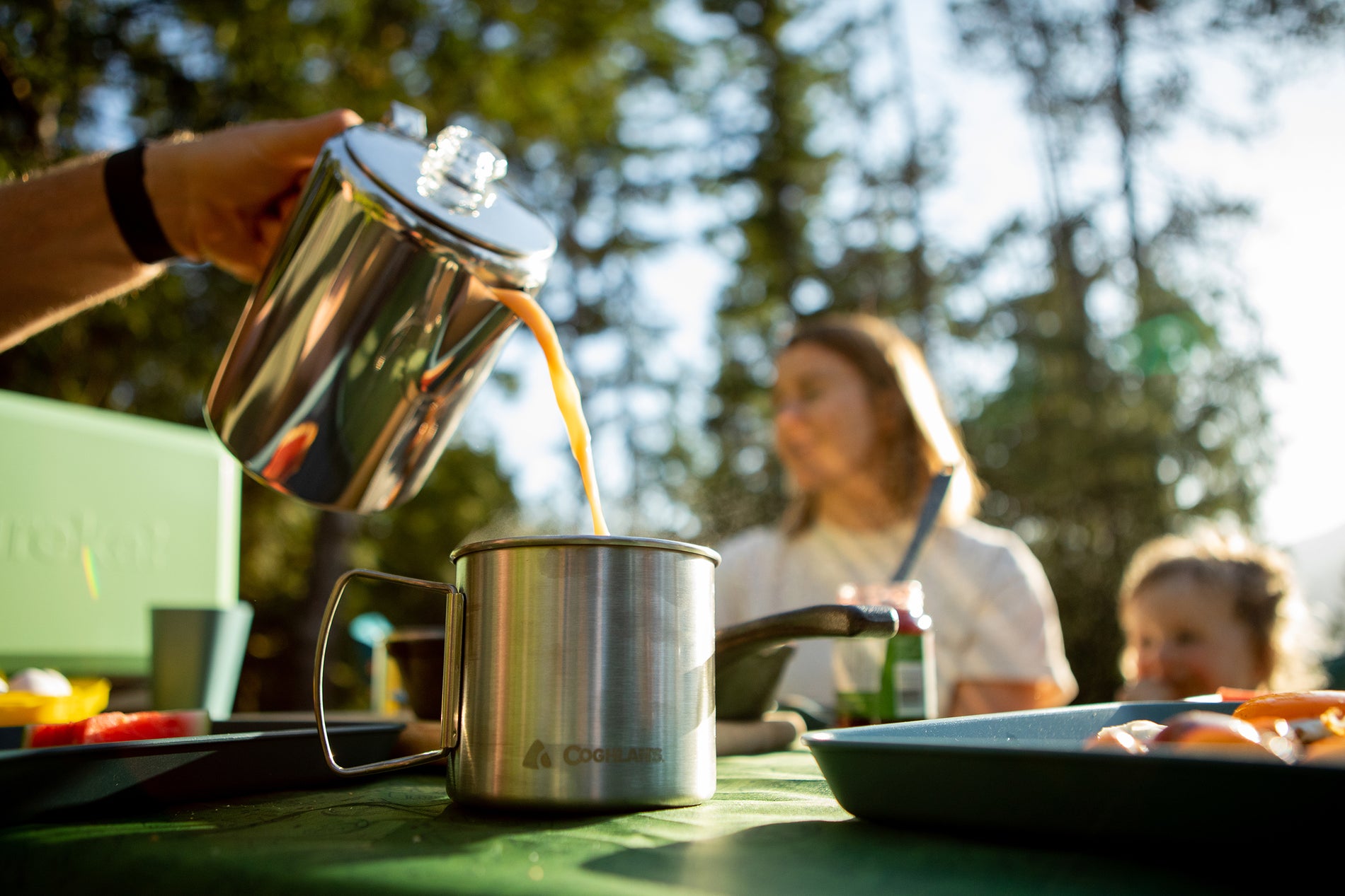 Mother and child enjoying fresh percolator coffee and breakfast together at a campsite while the father's hand pours coffee into a mug