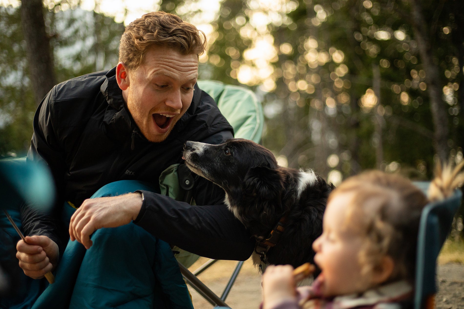 Black Collie sticking out it's tongue at it's owners, a dad and daughter, sitting in lawn chairs at a campsite 