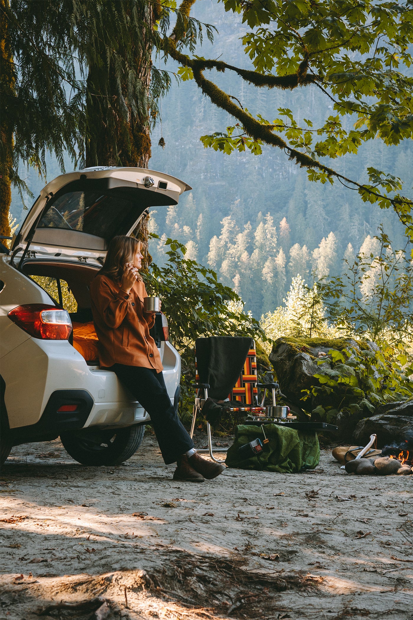 A young woman leans against an open car trunk at a campsite while enjoying her morning coffee