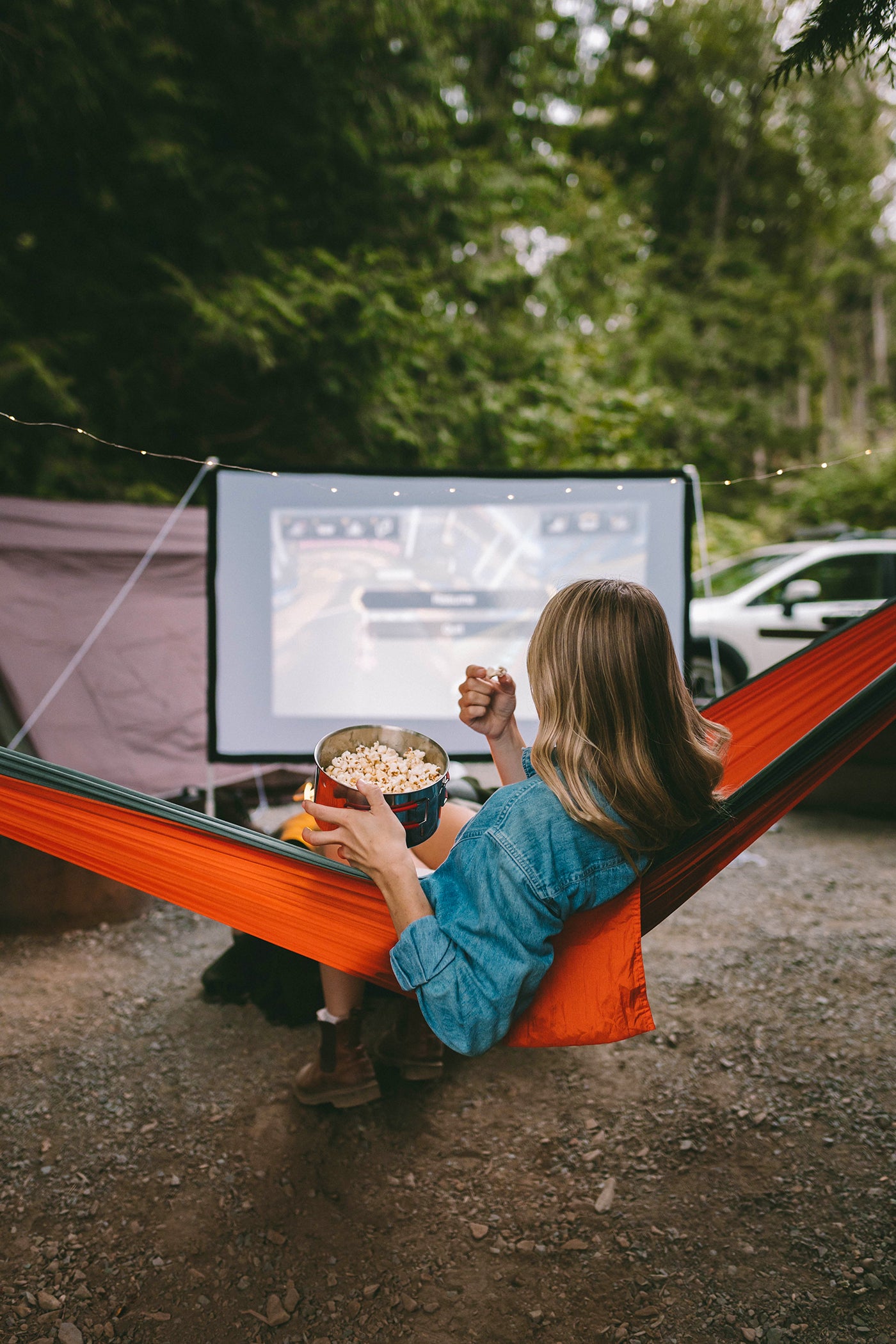 A young woman sitting in an orange hammock eating popcorn and facing an outdoor projector screen