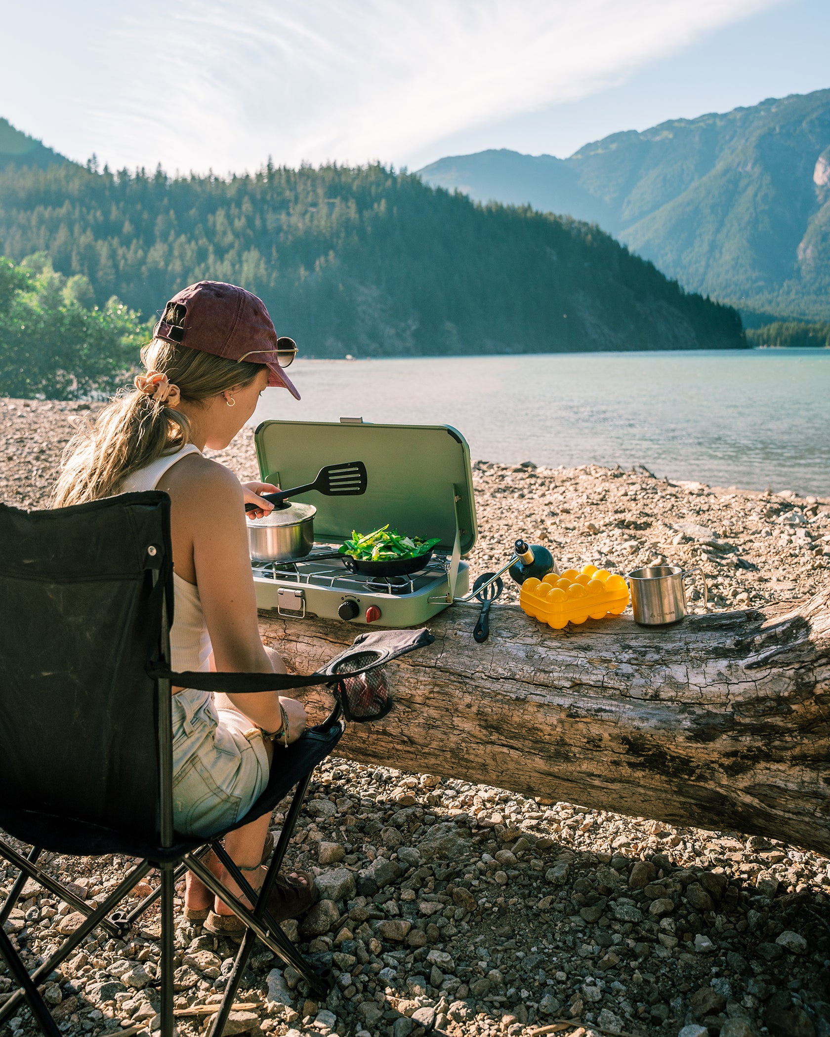 A young woman cooking on a portable propane camp stove on a log overlooking a lake and distant mountains